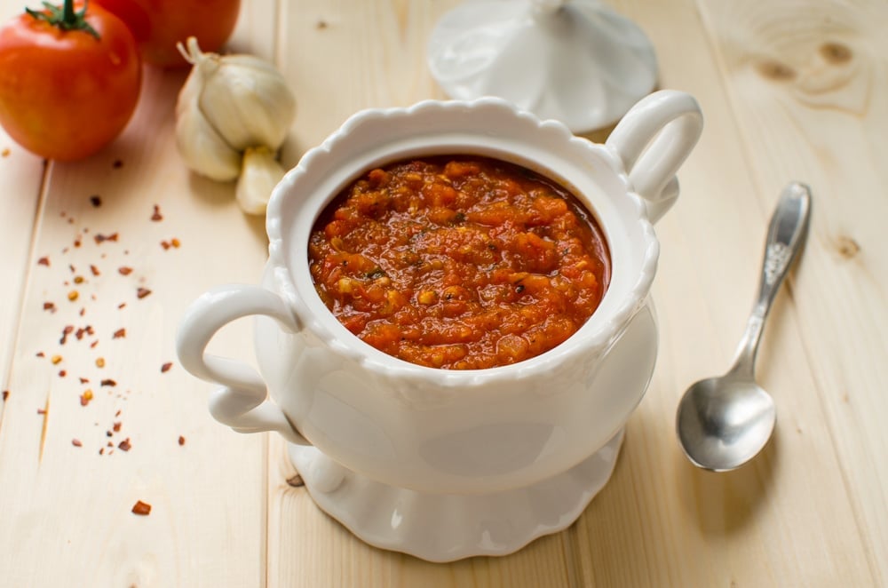 Homemade pizza sauce with fresh tomatoes in bowl with spoon on the side and raw garlic and tomato in the background.