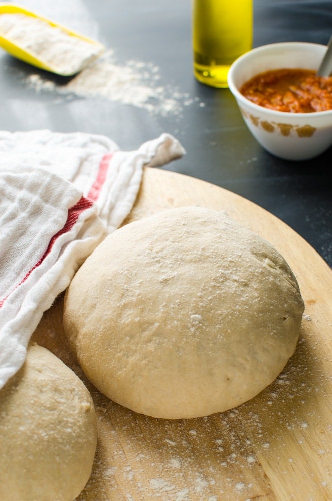 Homemade pizza dough balls on the rolling board with pizza sauce and some dry flour in the background.