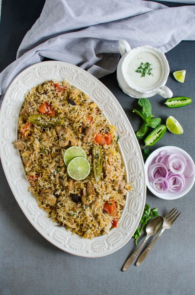 Top view of one pot chicken biryani rice in a large platter, garnished with sliced lime and served with sliced onion, yoghurt dipping on the side. 
