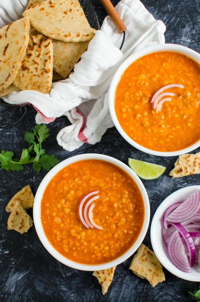 Red lentil curry in two serving bowl garnished with sliced onion and served with plain naan bread on the side. 