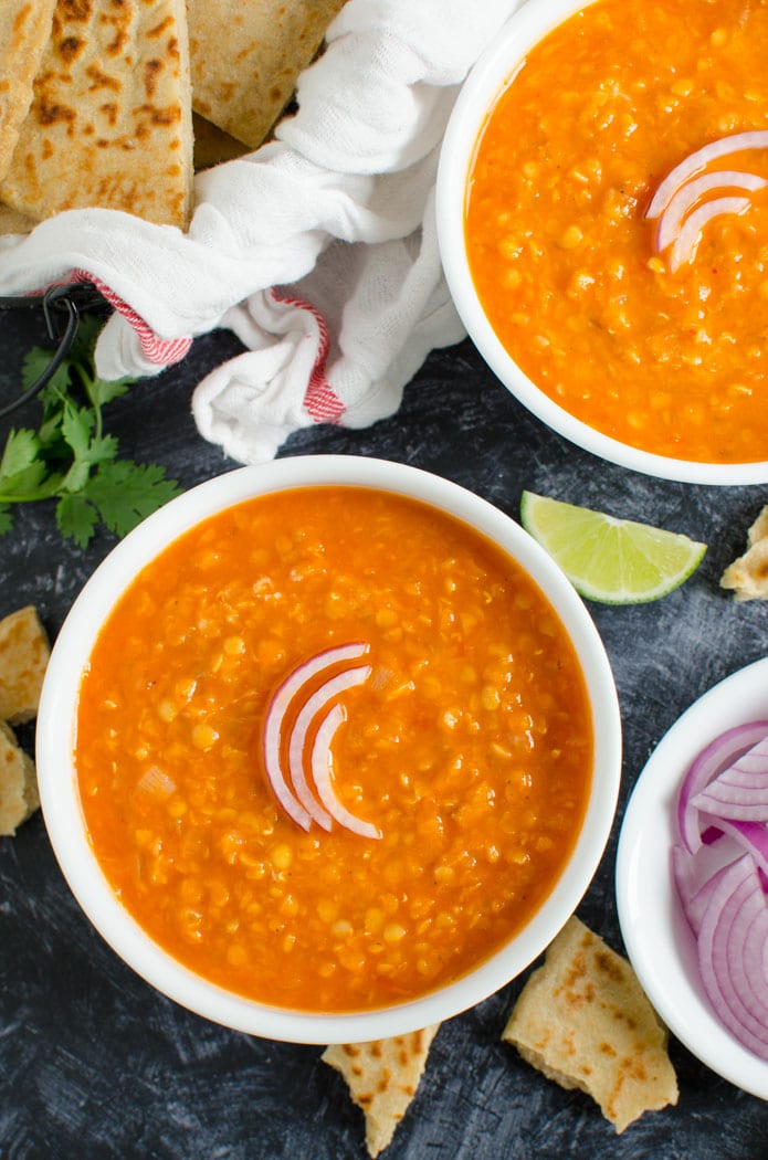 Red lentil curry in two serving bowl garnished with sliced onion and served with plain naan bread on the side. 