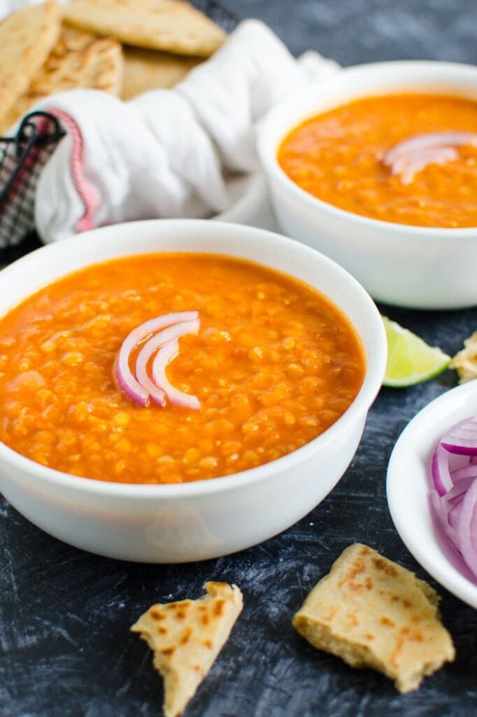 Red lentil curry in two serving bowl garnished with sliced onion and naan bread basket in the background.