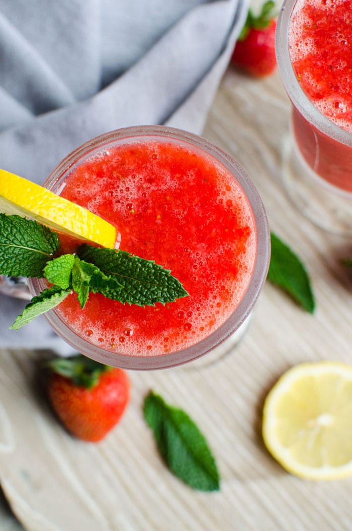 Strawberry slush in a serving glass with a paper straw, garnished with lemon slice, and fresh mint leaves.