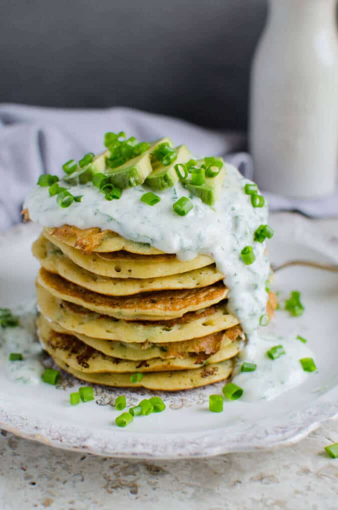 Stack of mung bean pancakes topped with yogurt sauce, slices of avocado and chopped scallions.