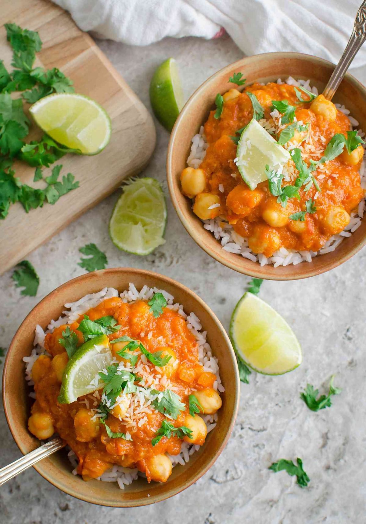 Coconut chickpea curry garnished with fresh cilantro and lime wedges in two serving bowls with metal spoon.