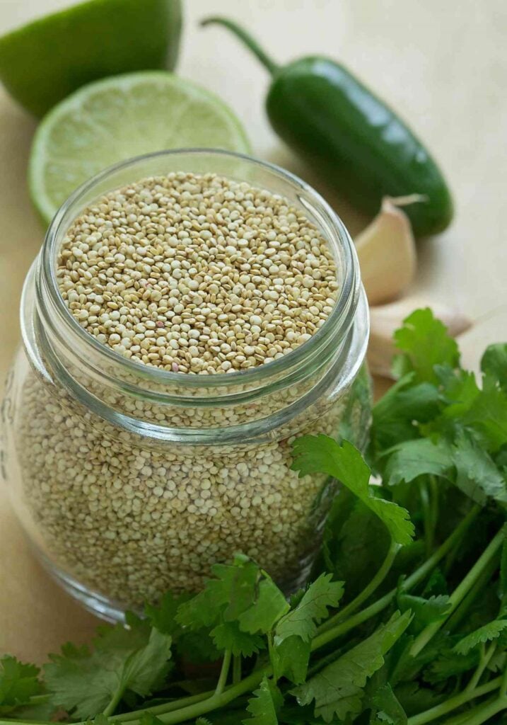 Raw quinoa in a glass jar, fresh cilantro. garlic, jalapeño, and lime placed on a flat surface.