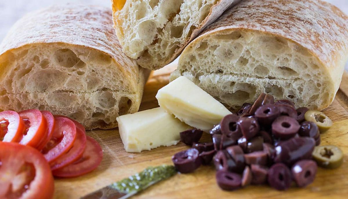 Sandwich bread, cheese, tomato slices and olives on a lat surface.