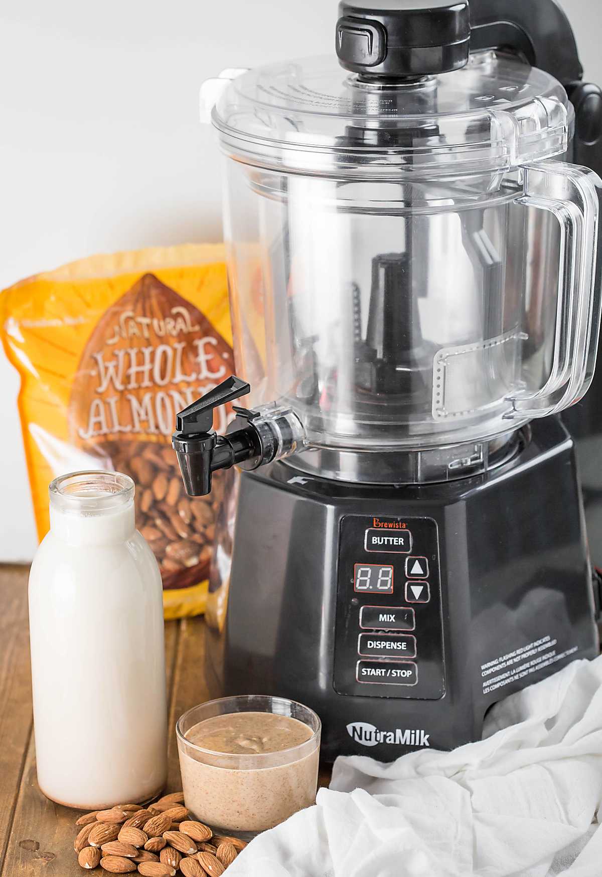 The Nutramilk machine with almond milk in glass bottle, almond butter in glass bowl and some almonds.