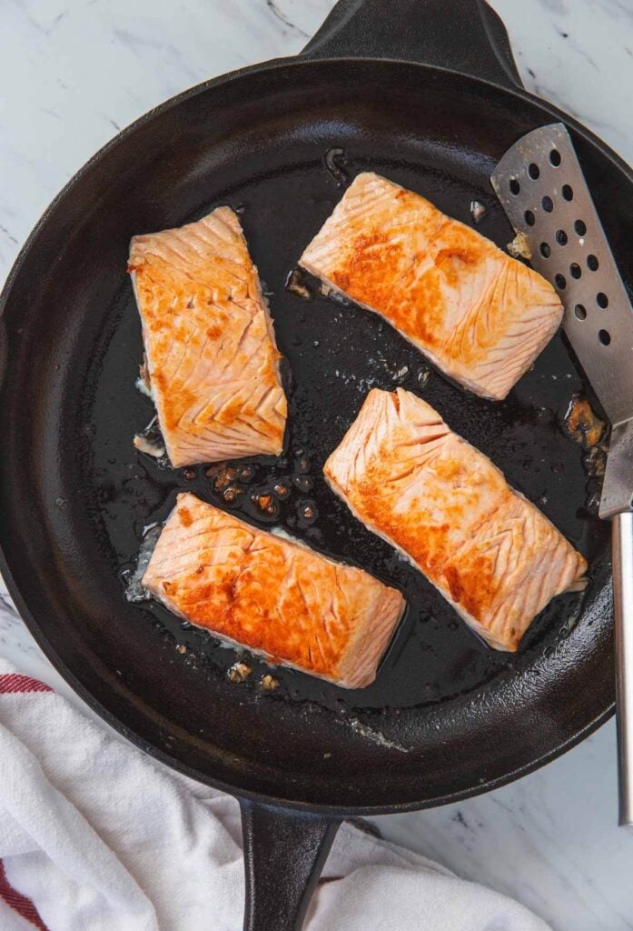 process of searing salmon fillets in a large cast iron skillets with stainless steel spatula.