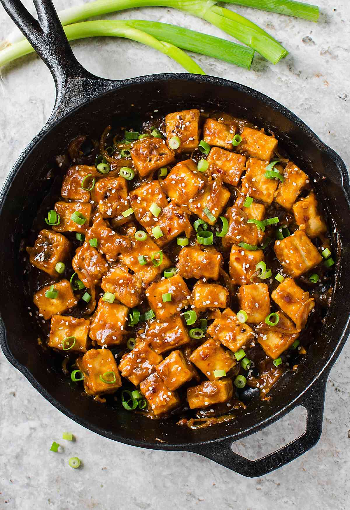 Asian chili garlic tofu garnished with fresh spring onions in a black cast iron pan.