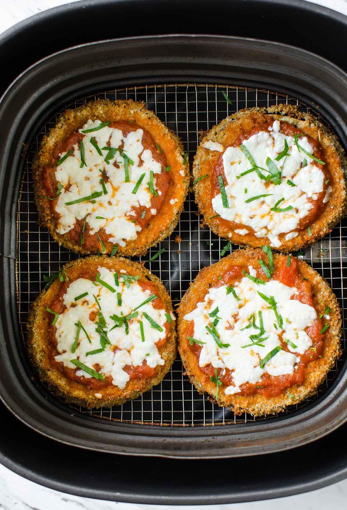 Air fried eggplant parmesan garnished with fresh basil in a air fryer basket.