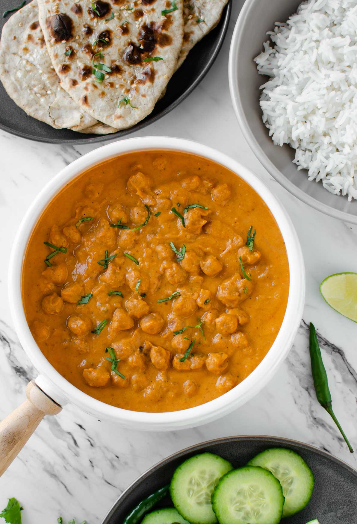Chickpea tikka masala garnished with fresh cilantro in a serving bowl and served with whole wheat naan, plain rice, and cucumber slices on the side.