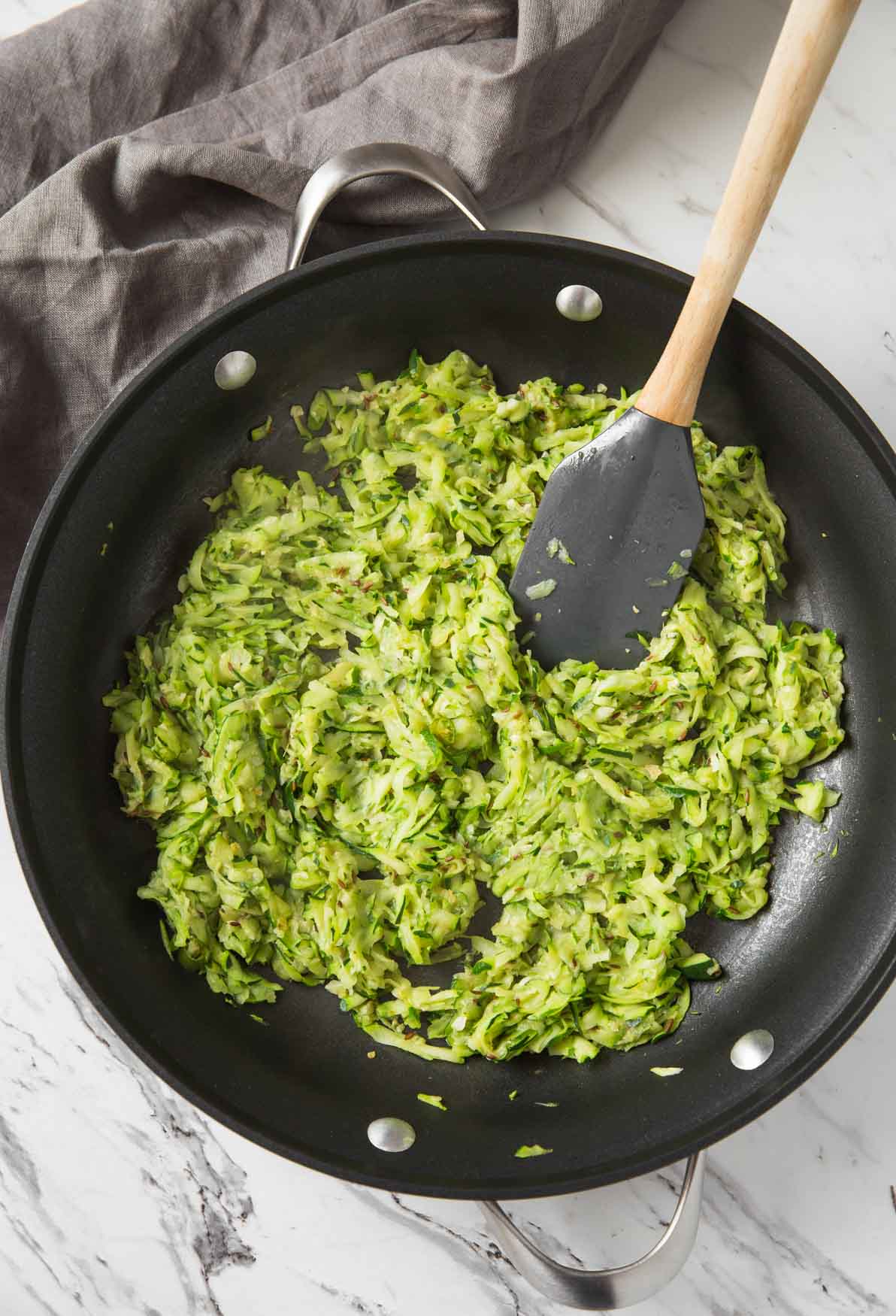 Sautéing grated zucchini in a large frying pan with a rubber spatula.