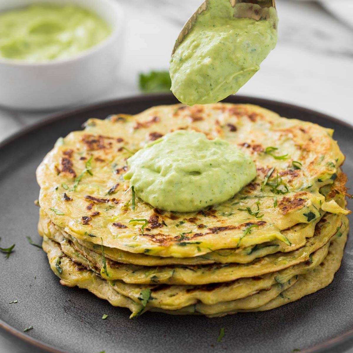 Healthy zucchini pancakes stacked in a serving plate with a dollop of green avocado sauce.