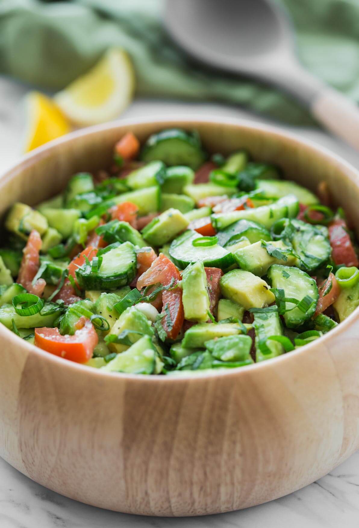 photo of avocado cucumber salad in a large wooden salad bowl and serving spoon and lemon wedges on the side.