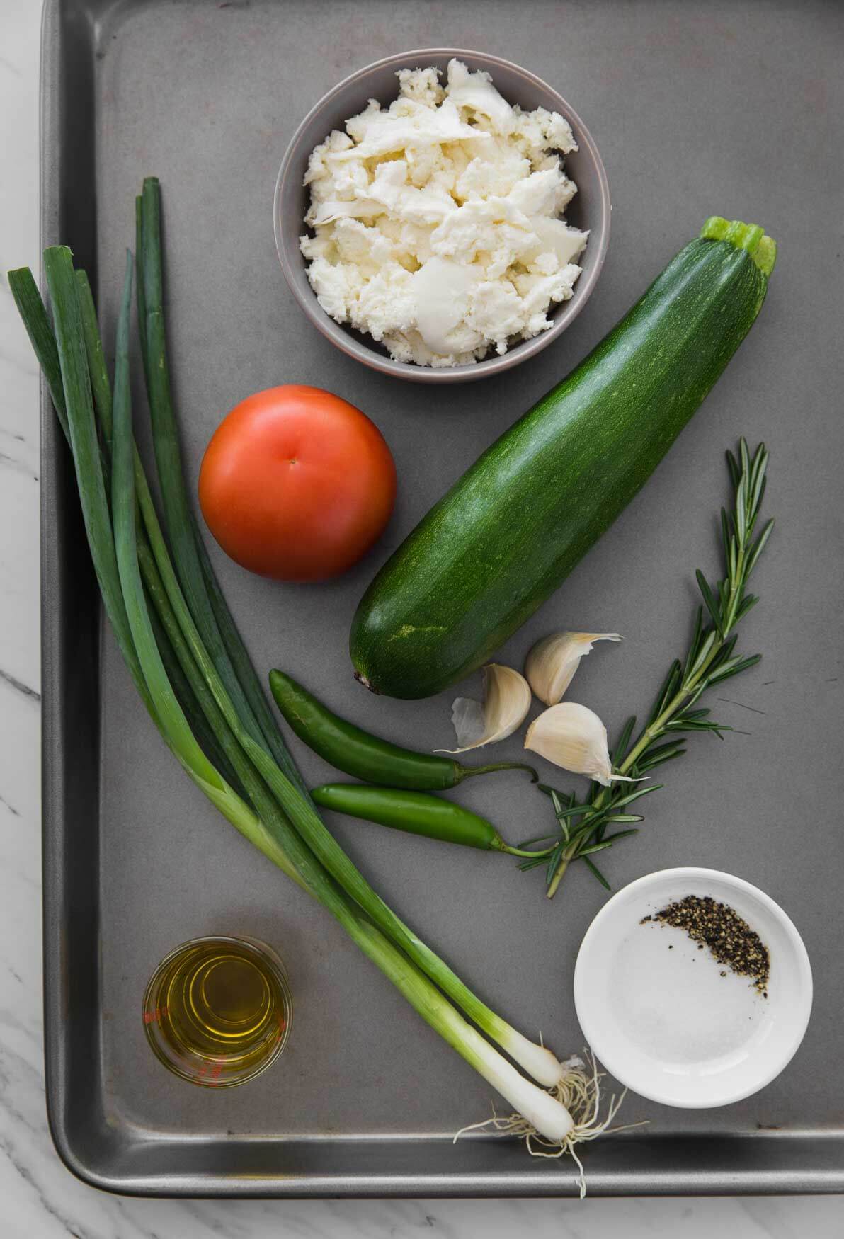 image of ingredients for making flatbread. It includes fresh zucchini, scallions, cheese, tomato, whole garlic, oil, rosemary, salt etc.