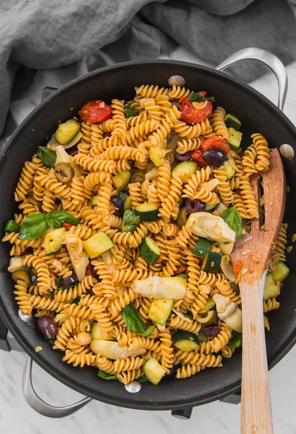 Healthy Mediterranean pasta with artichokes and olives in a large skillet with a wooden spatula.