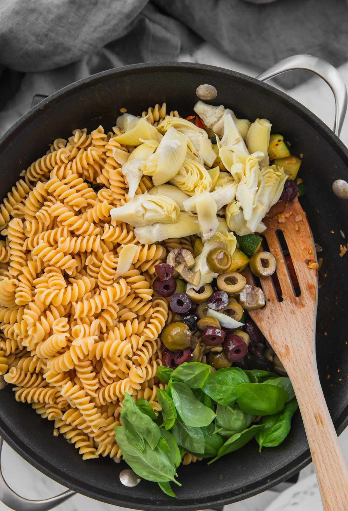 Healthy Mediterranean pasta with artichokes and olives while mixing in a large skillet with a wooden spatula.