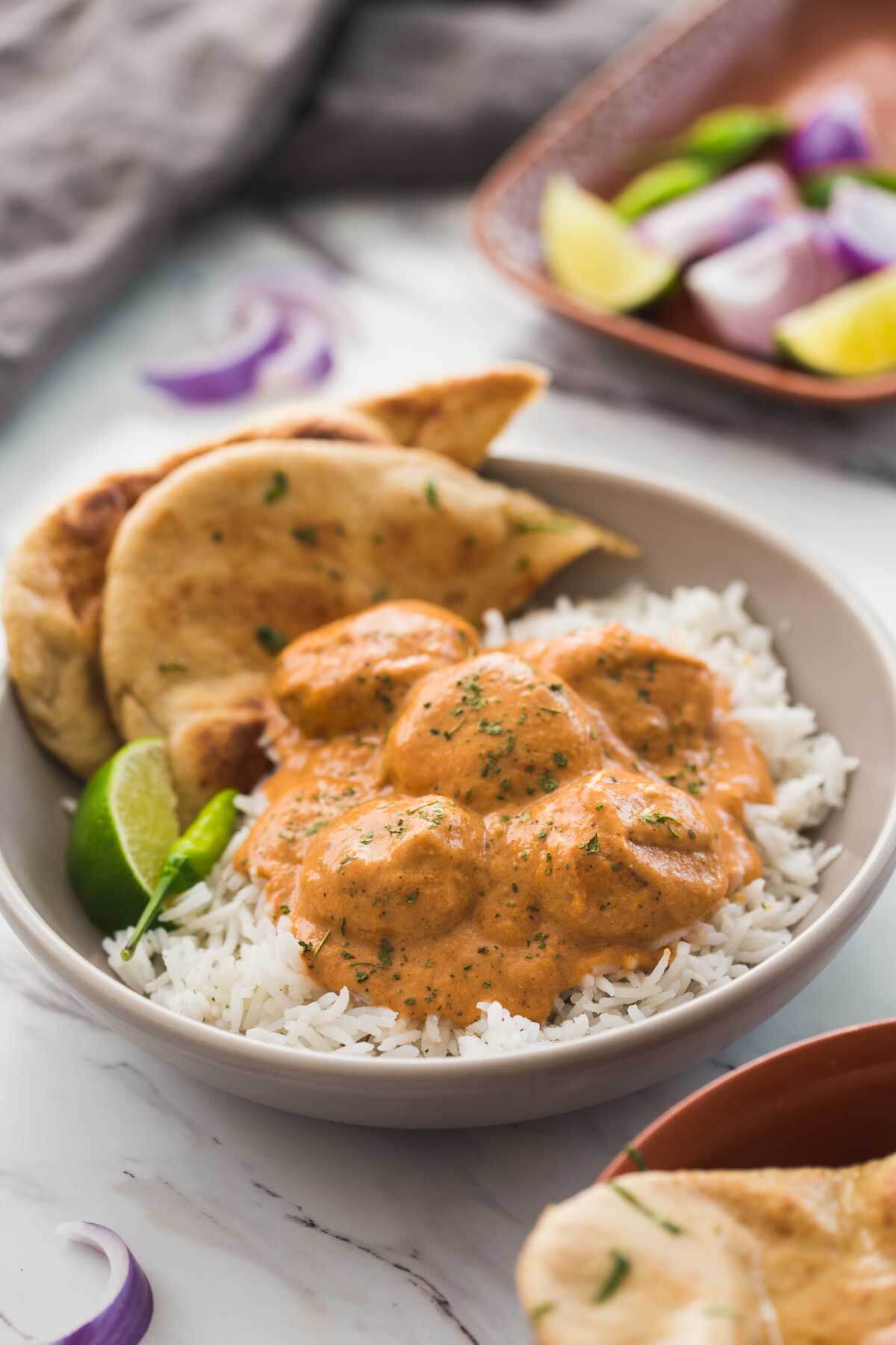 Butter chicken meatballs in a serving dish with naan bread, plain rice, lime wedge and green chili on the side.