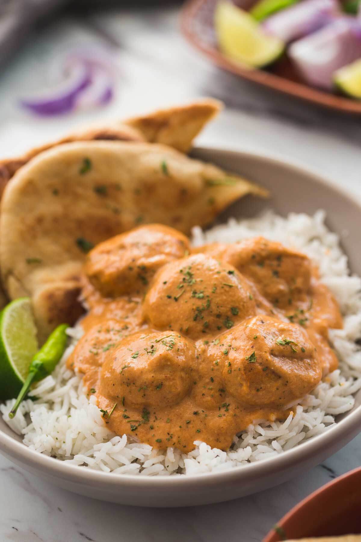 Butter chicken meatballs in a serving dish with naan bread, plain rice, lime wedge and green chili on the side.