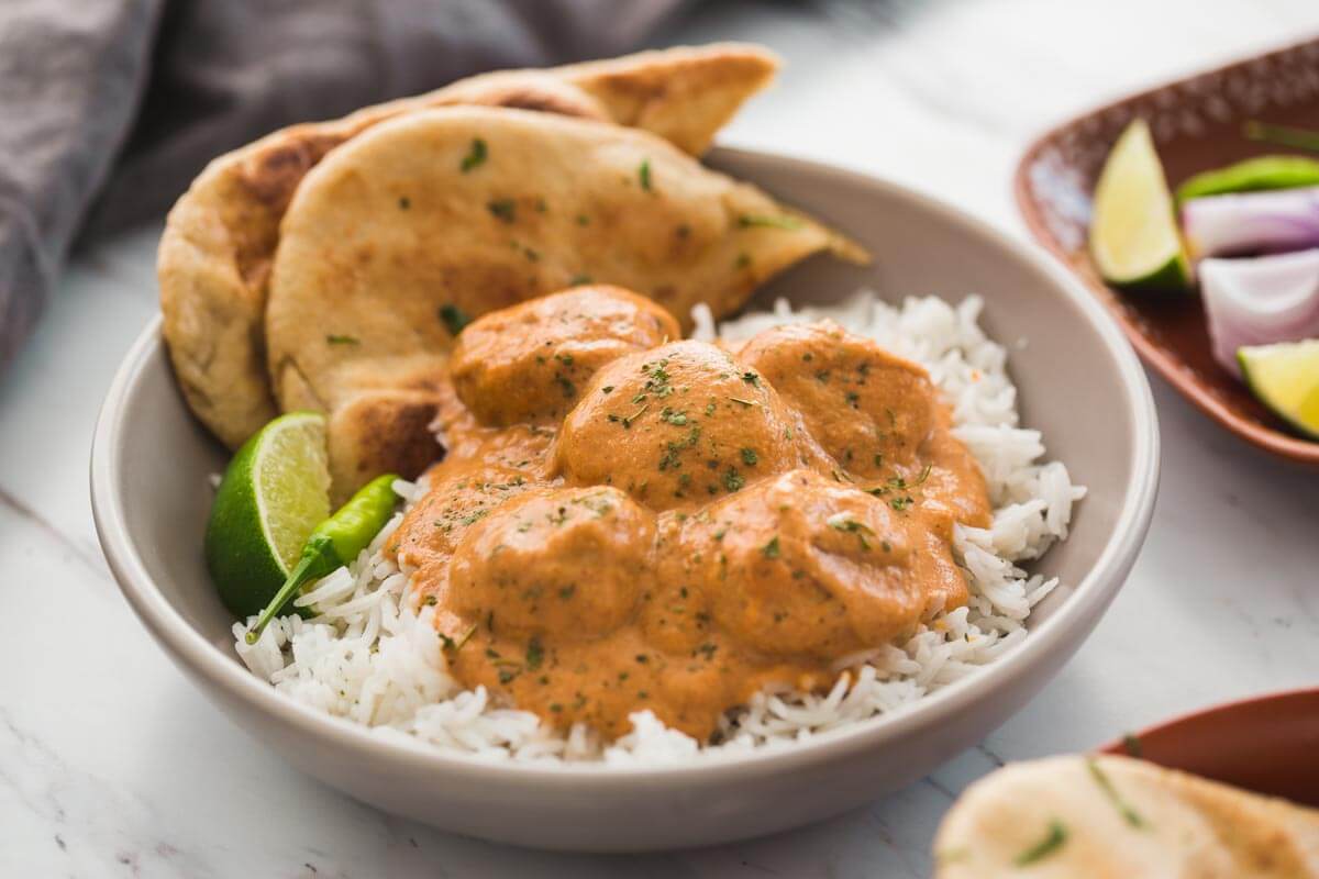 Butter chicken meatballs in a serving dish with naan bread, plain rice, lime wedge and green chili on the side.