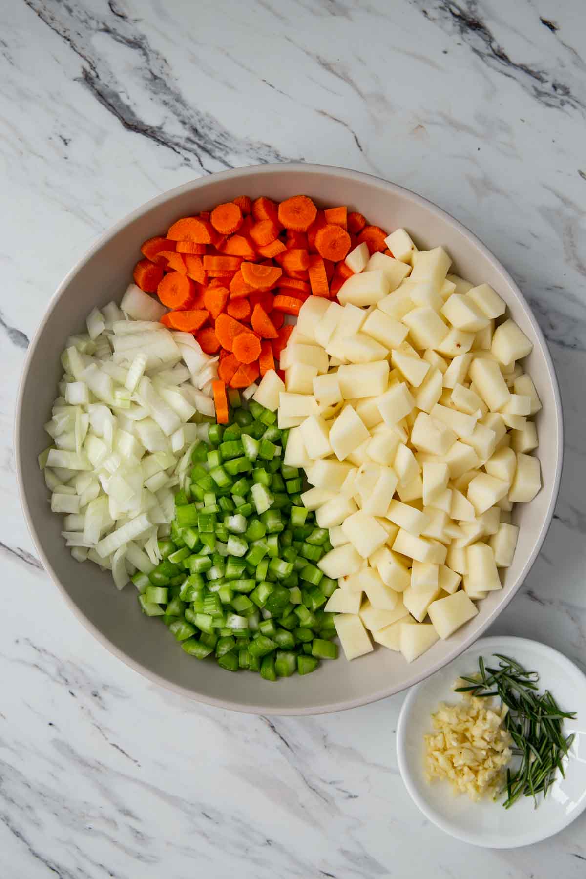 Diced potato, celery, onion, and carrot in a large bowl along with chopped garlic and fresh rosemary leaves in a small dish.