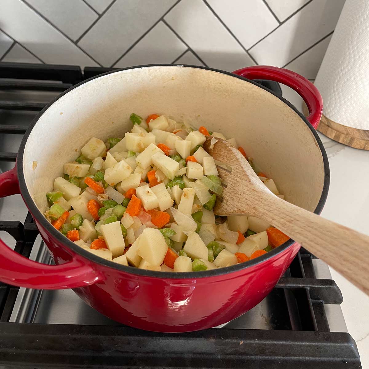 Sautéing diced potato, carrot, and celery in a dutch oven with a wooden spatula on a gas stove.