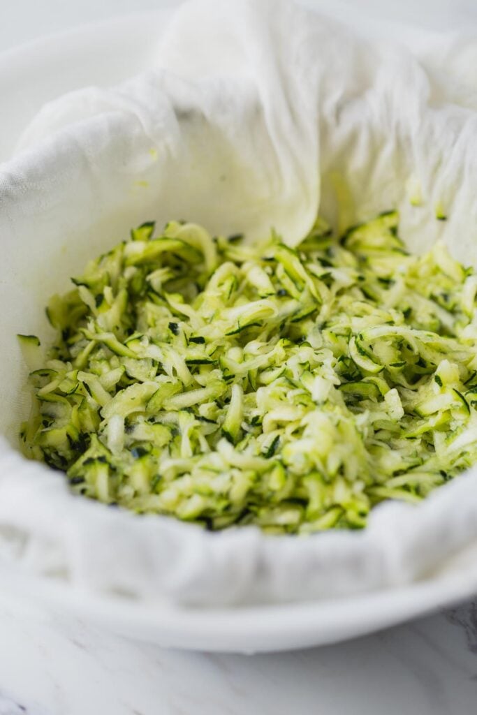 Shredded zucchini in a bowl lined with cheesecloth. 