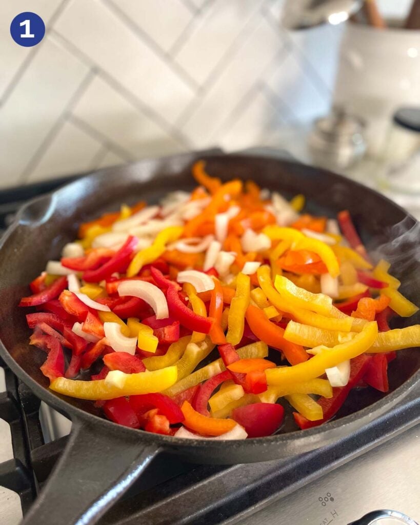 Sliced peppers and onion in a cast iron pan on a gas stove.