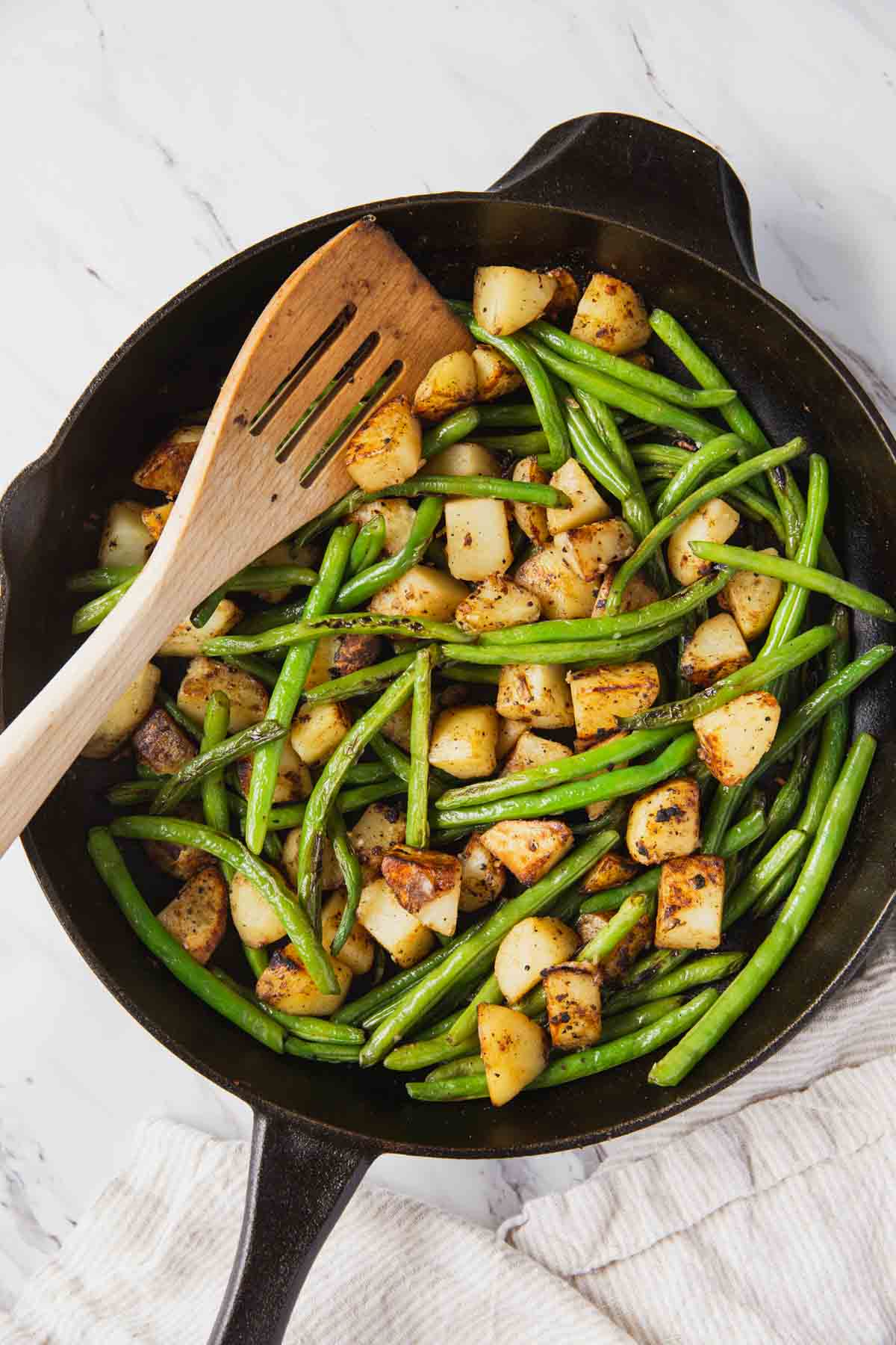 Roasted green beans and potatoes in a cast iron pan with a wooden spatula.