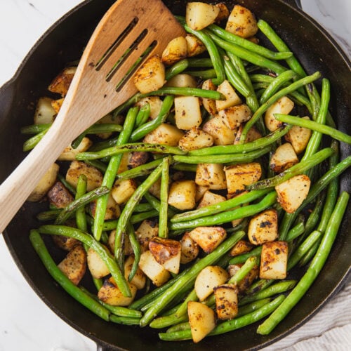 Roasted green beans and potatoes in a cast iron pan with wooden spatula.