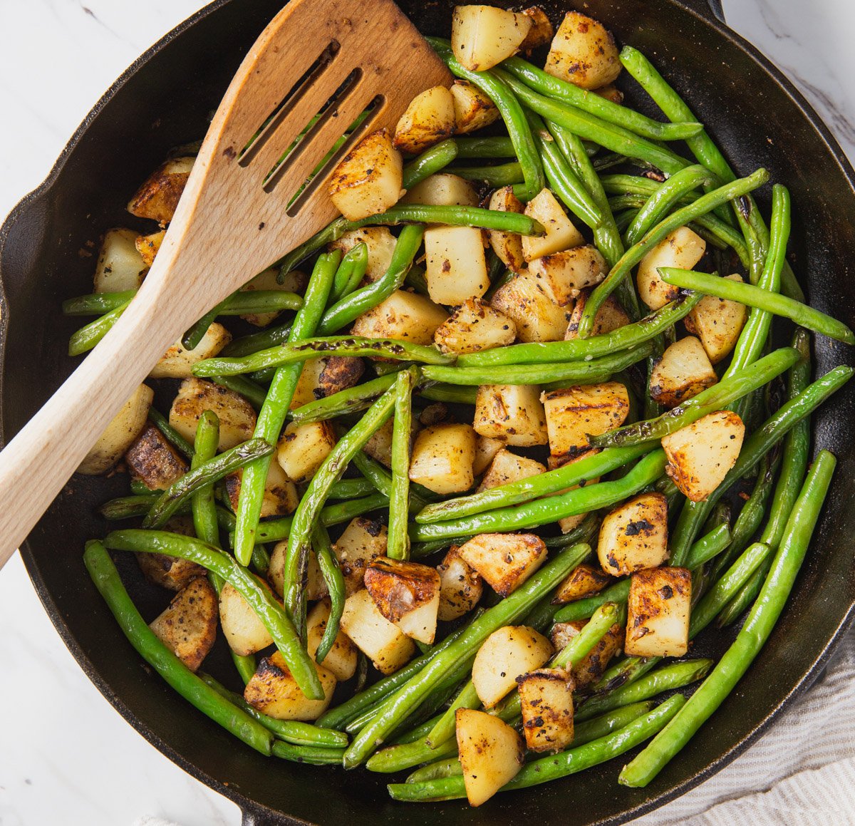 Roasted green beans and potatoes in a cast iron pan with wooden spatula.