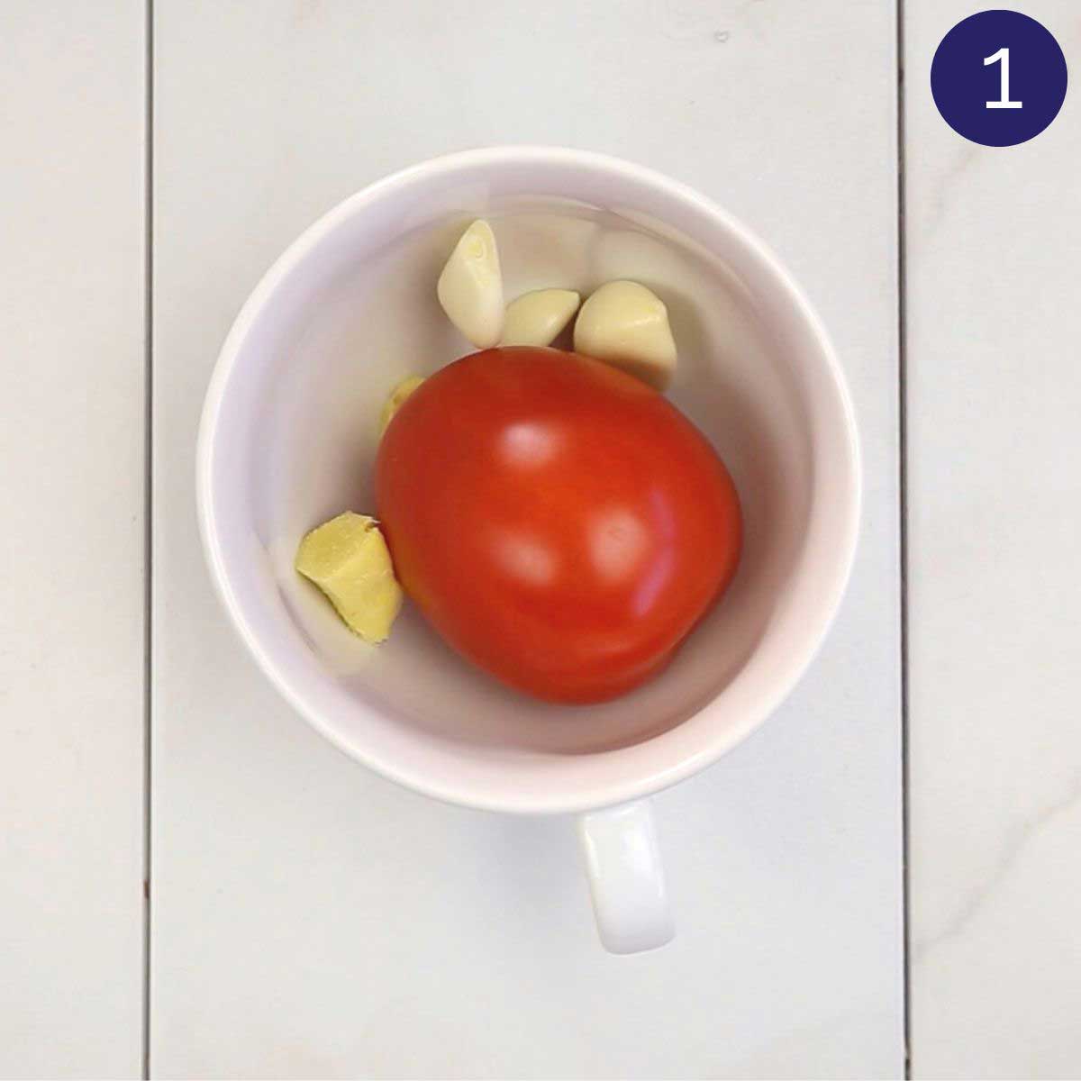 One red tomato, garlic cloves and ginger pieces in a mixing cup.