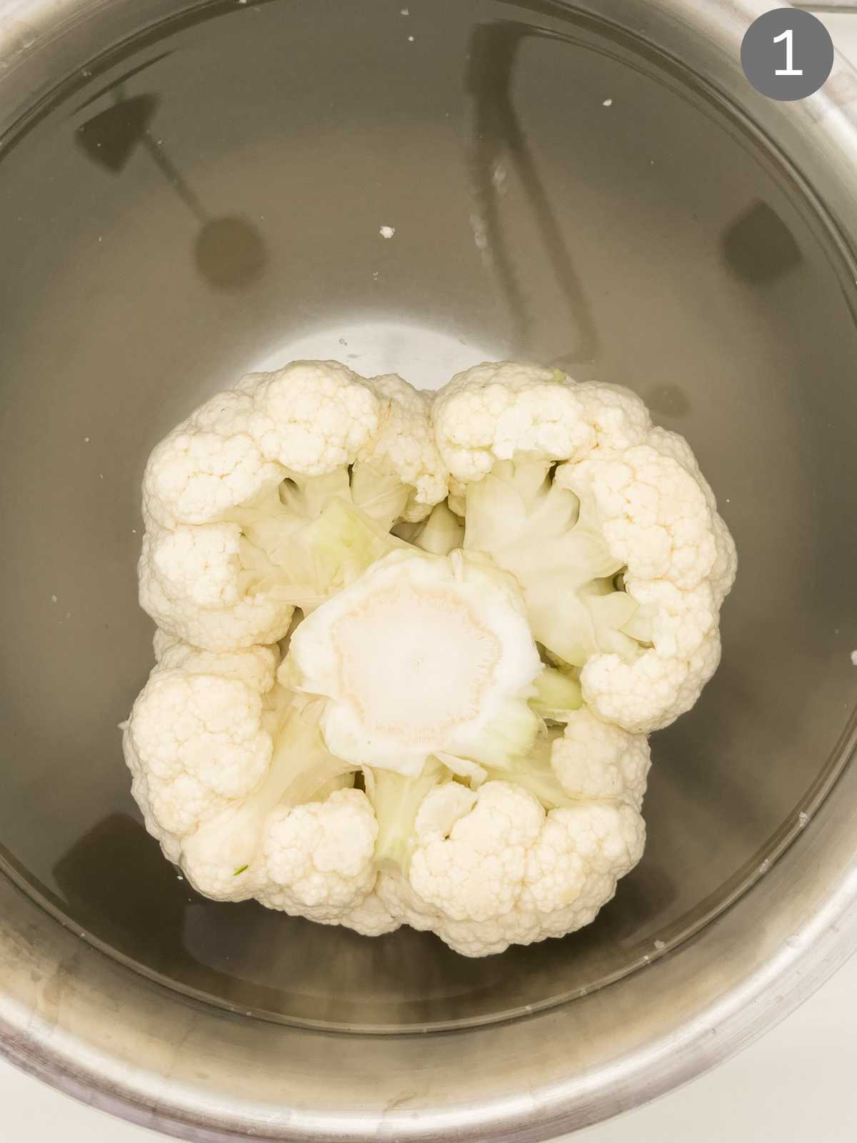 Whole cauliflower head submerged in water in a mixing bowl. 