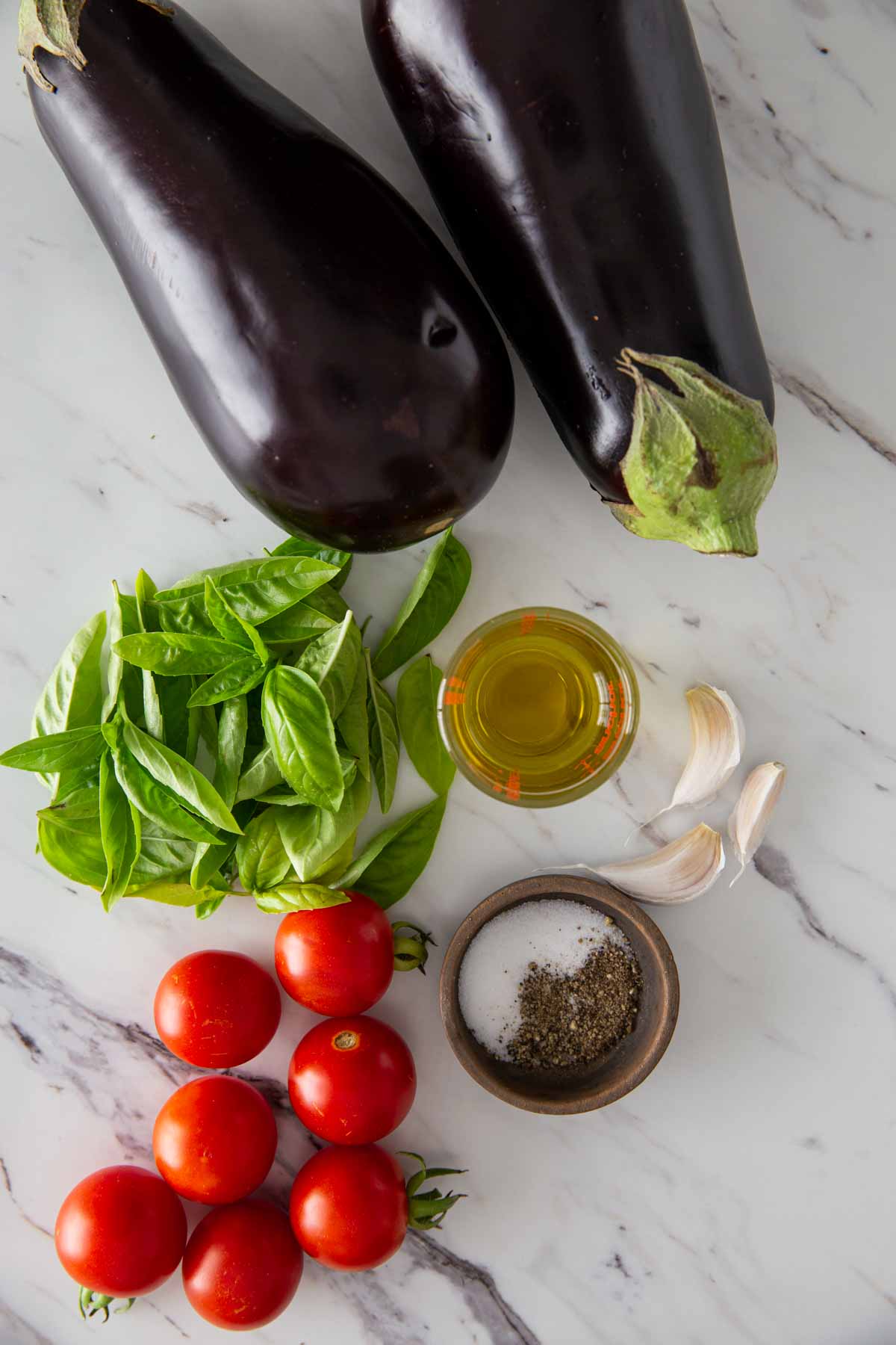 Fresh ingredients, such as eggplant, cherry tomatoes, garlic, basil, seasonings, and oil, are laid out on a flat surface.