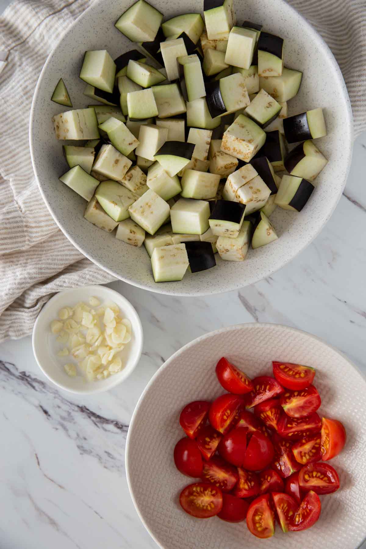 Diced eggplant in one bowl, cherry tomatoes cut into quarters in another bowl, and freshly chopped garlic in a small bowl for making the healthy eggplant recipe.