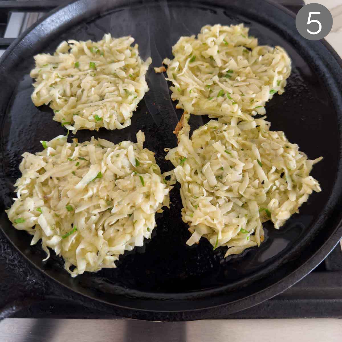 Four potato fritters on a cast iron pan after cooking for some time and before flipping.