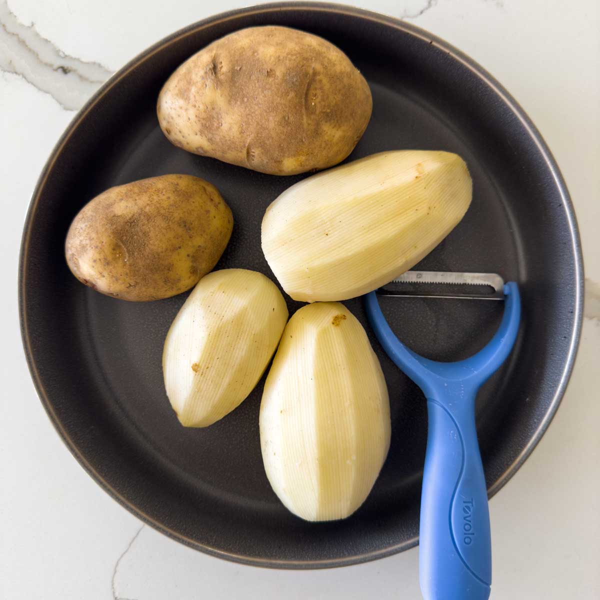 Some peeled and unpeeled potatoes with a peeler in a large dish.