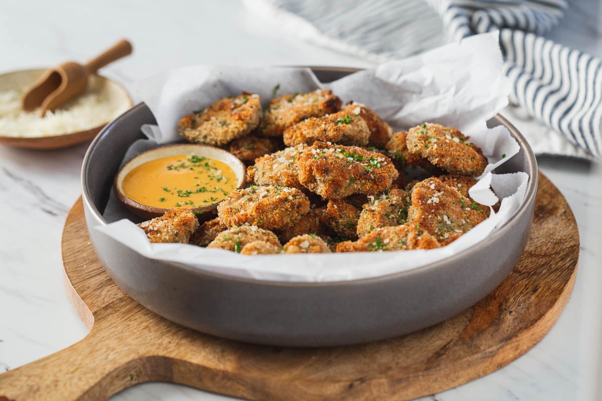 Breaded ground chicken nuggets in a serving shallow bowl with the dipping sauce on the side are ready to serve. The bowl is placed over a round wooden board.