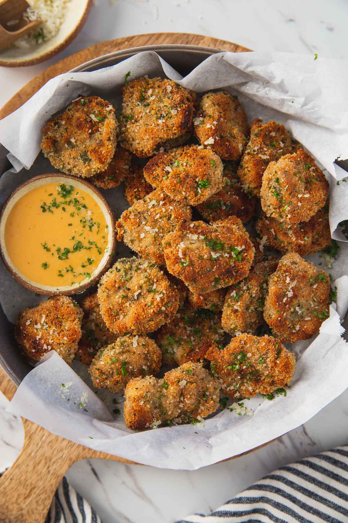 Breaded ground chicken nuggets in a serving shallow bowl with the dipping sauce on the side are ready to serve. The bowl is placed over a round wooden board.