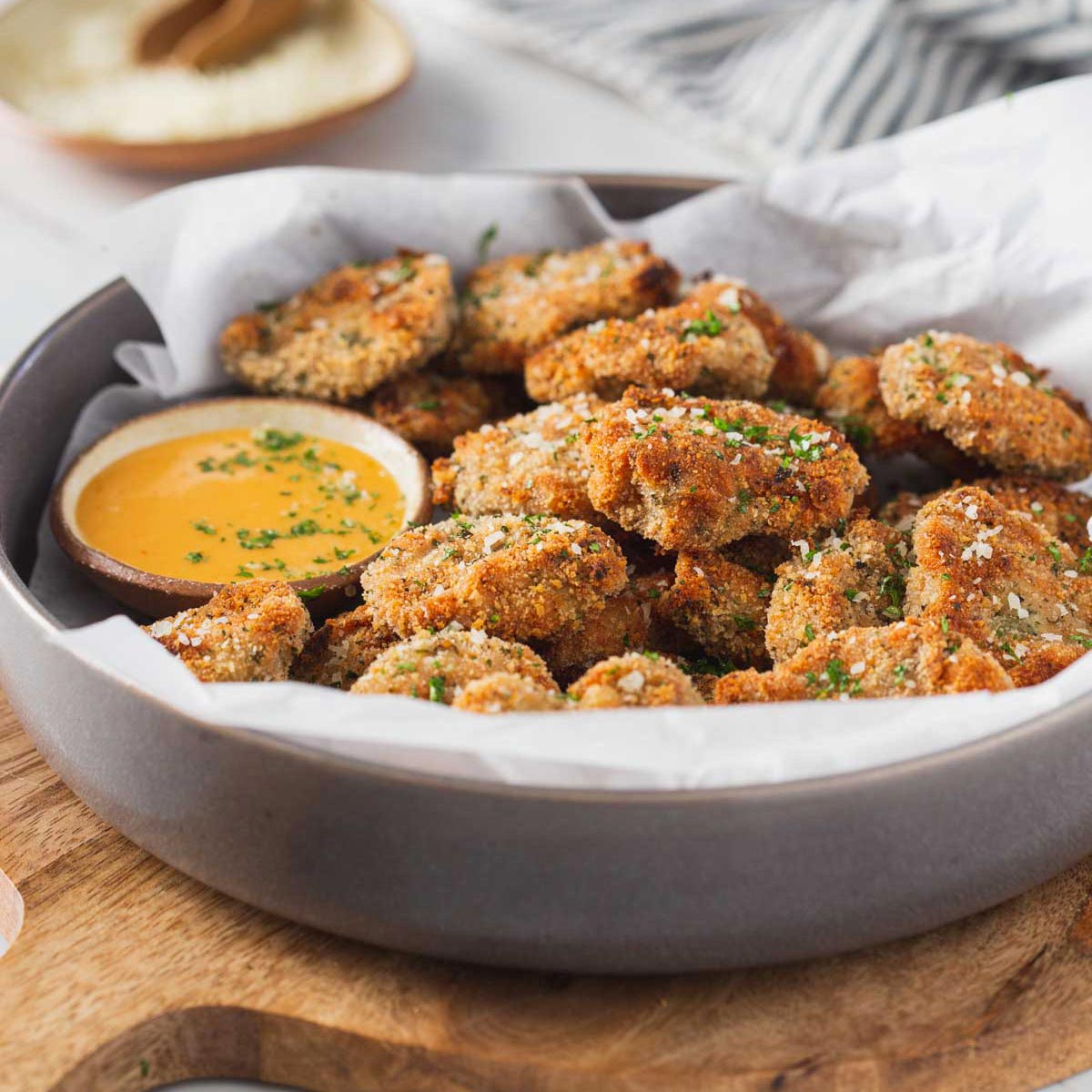 Breaded ground chicken nuggets in a serving shallow bowl with the dipping sauce on the side are ready to serve. The bowl is placed over a round wooden board.