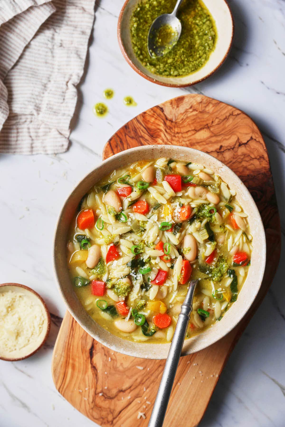 White beans, pesto, and vegetable orzo soup are served in a bowl with a metal spoon on a wooden riser and some parmesan cheese and pesto in mini dishes on the side. 