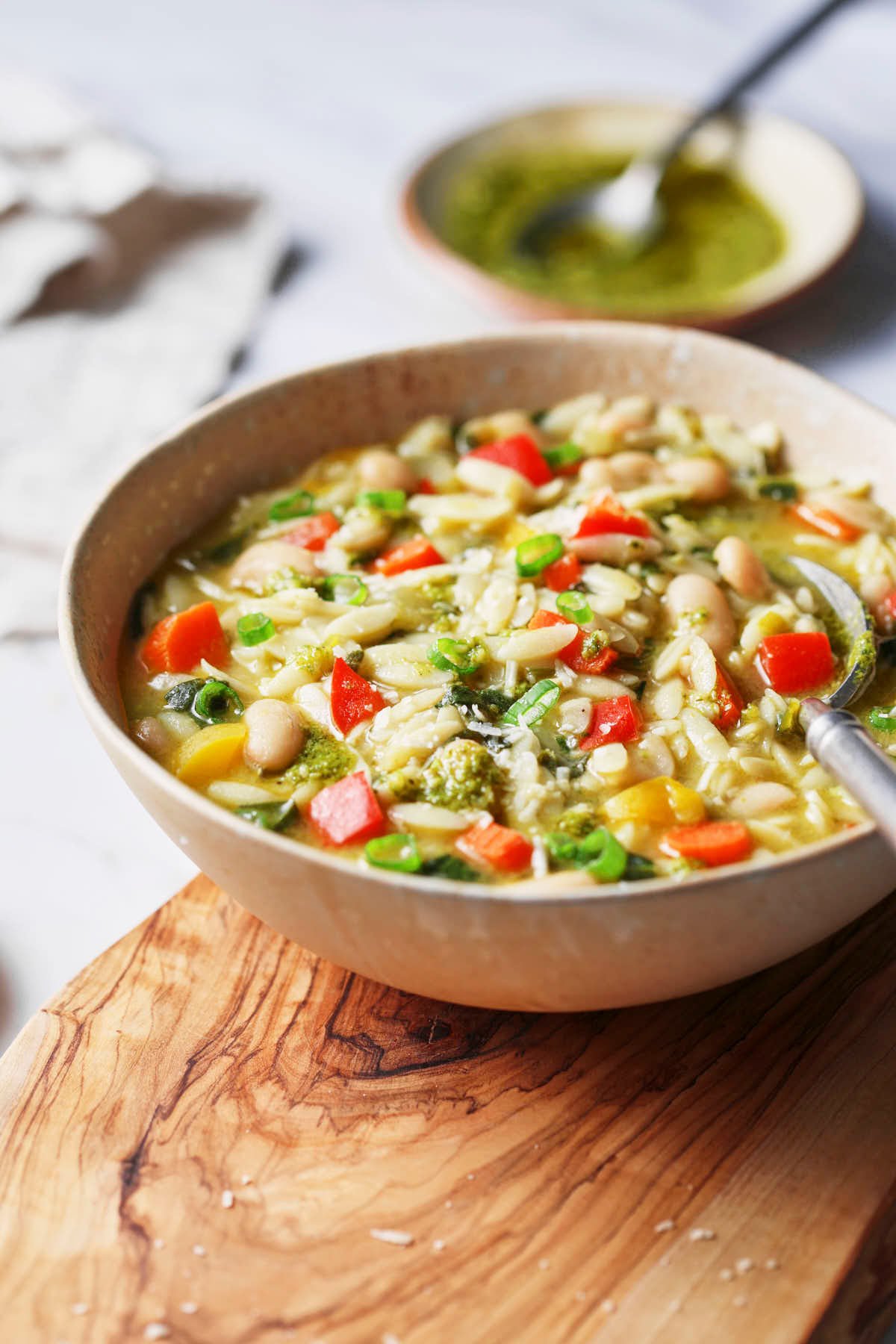 White beans, pesto, and veggie orzo soup are served in a bowl with a metal spoon on a wooden riser and some basil pesto in a mini dish on the side. 