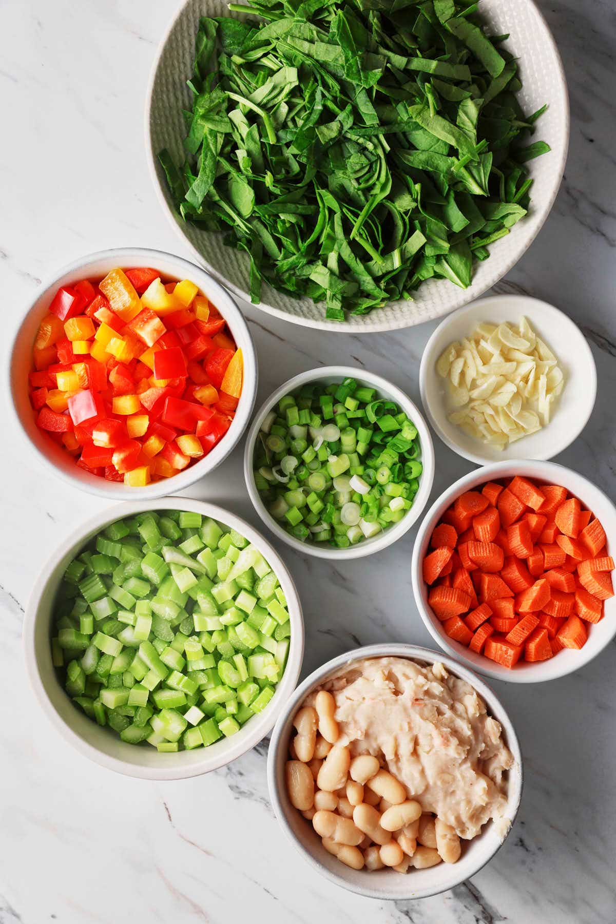 Diced vegetables, sliced garlic, and smashed and whole cannellini beans are placed in different white bowls on a flat surface.