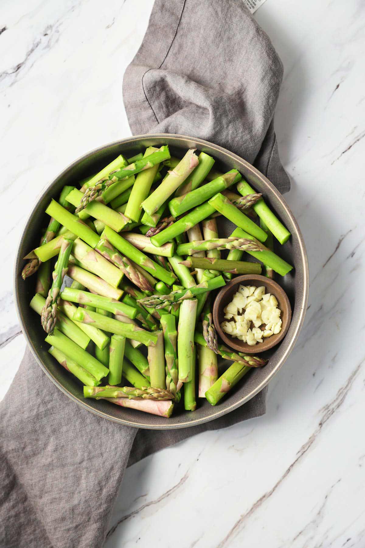 Asparagus cut into 2" pieces and placed in a shallow dish with sliced garlic in a condiment bowl. The dish is placed on a gray napkin on a flat surface. 