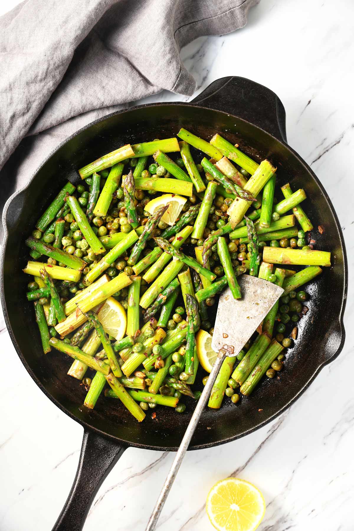 Roasted asparagus and peas in a cast iron pan with a metal spatula. On the side, there is a gray napkin and a lemon piece. 
