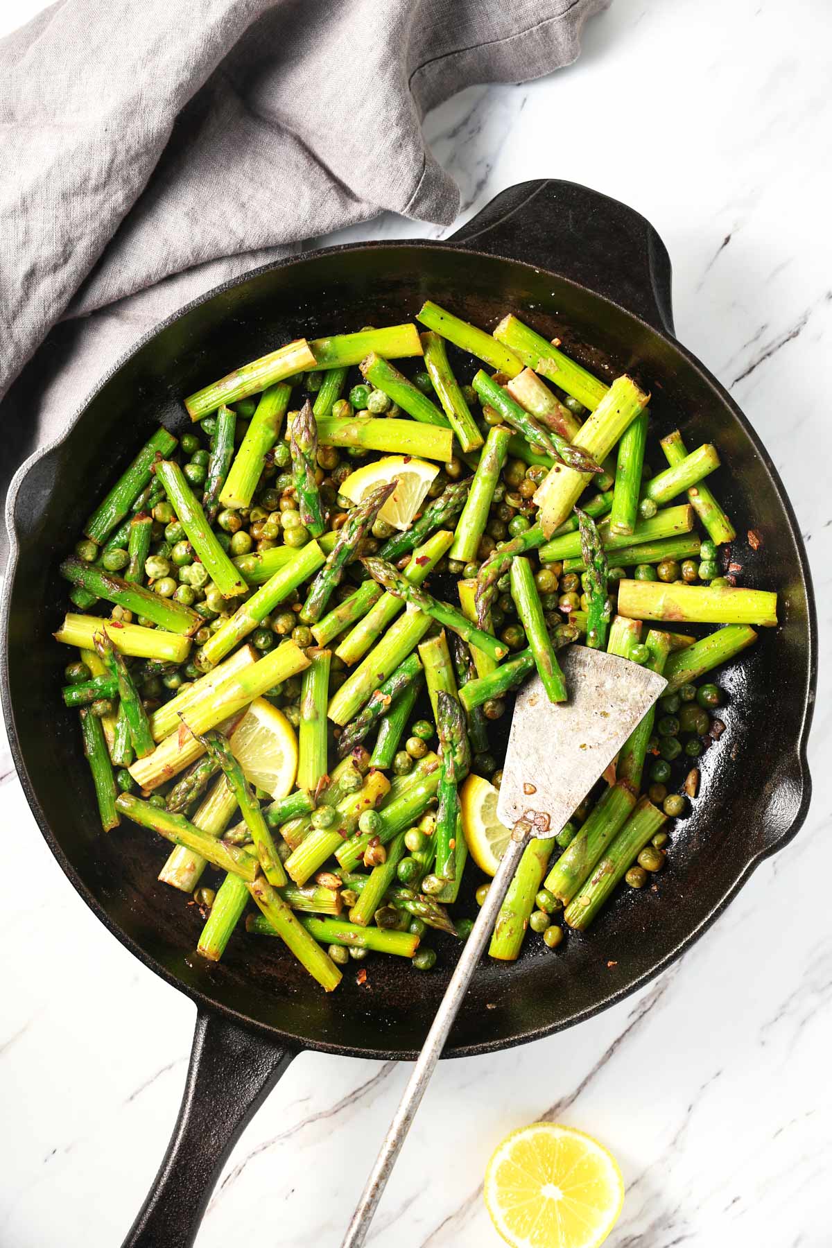 Roasted asparagus and peas in a cast iron pan with a metal spatula. On the side, there is a gray napkin and a lemon piece.