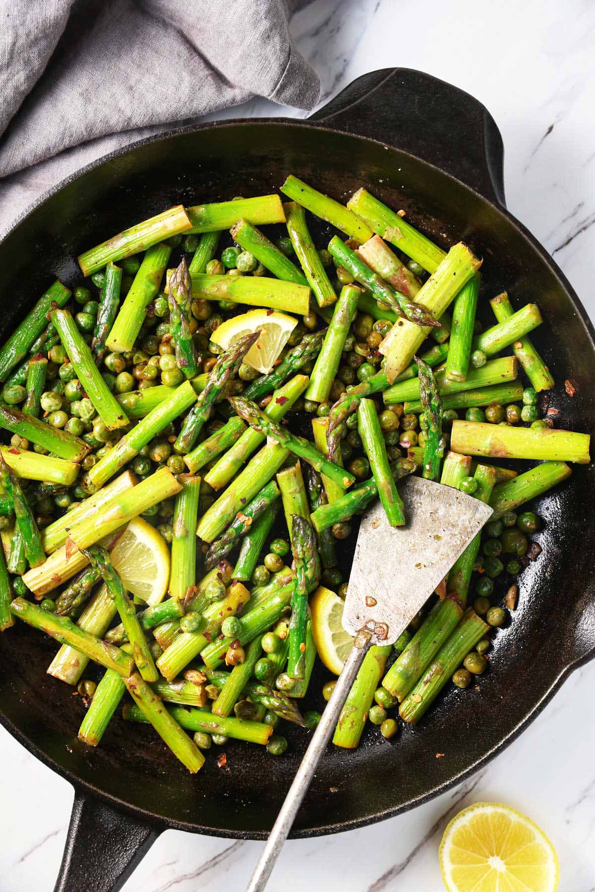 Roasted asparagus and peas in a cast iron pan with a metal spatula. There is a gray napkin and a half-lemon piece on the side. 