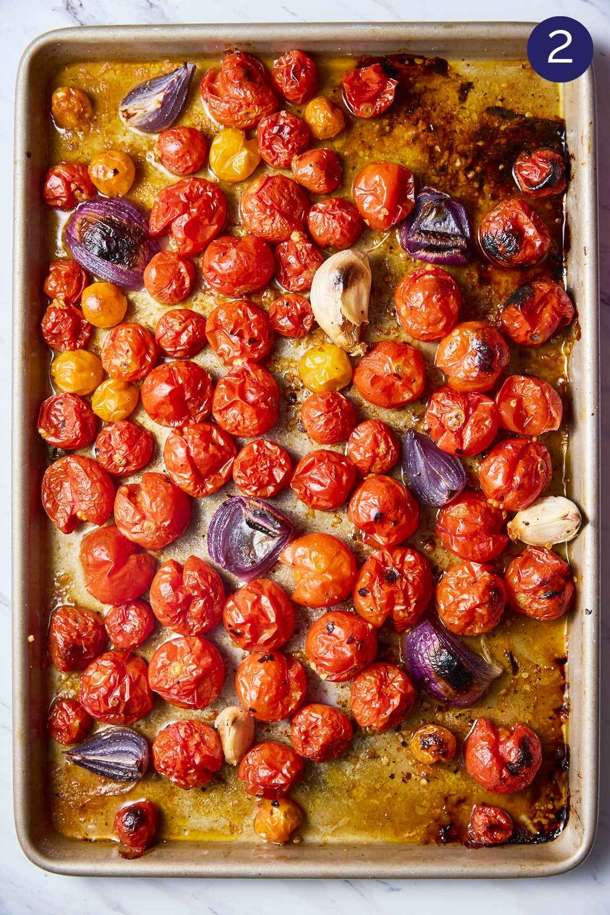 Cherry tomatoes with onion, garlic cloves and seasonings on baking sheet after roasting.