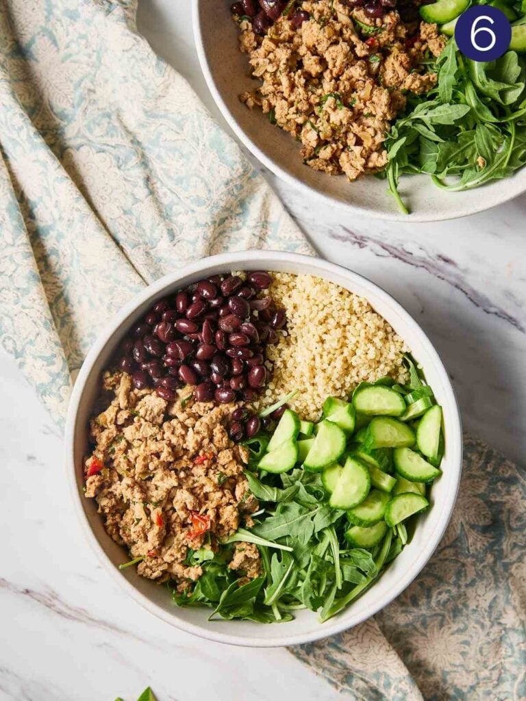 Cooked quinoa, fresh arugula and cooked ground turkey, black beans and sliced cucumber arranged in a serving bowl.